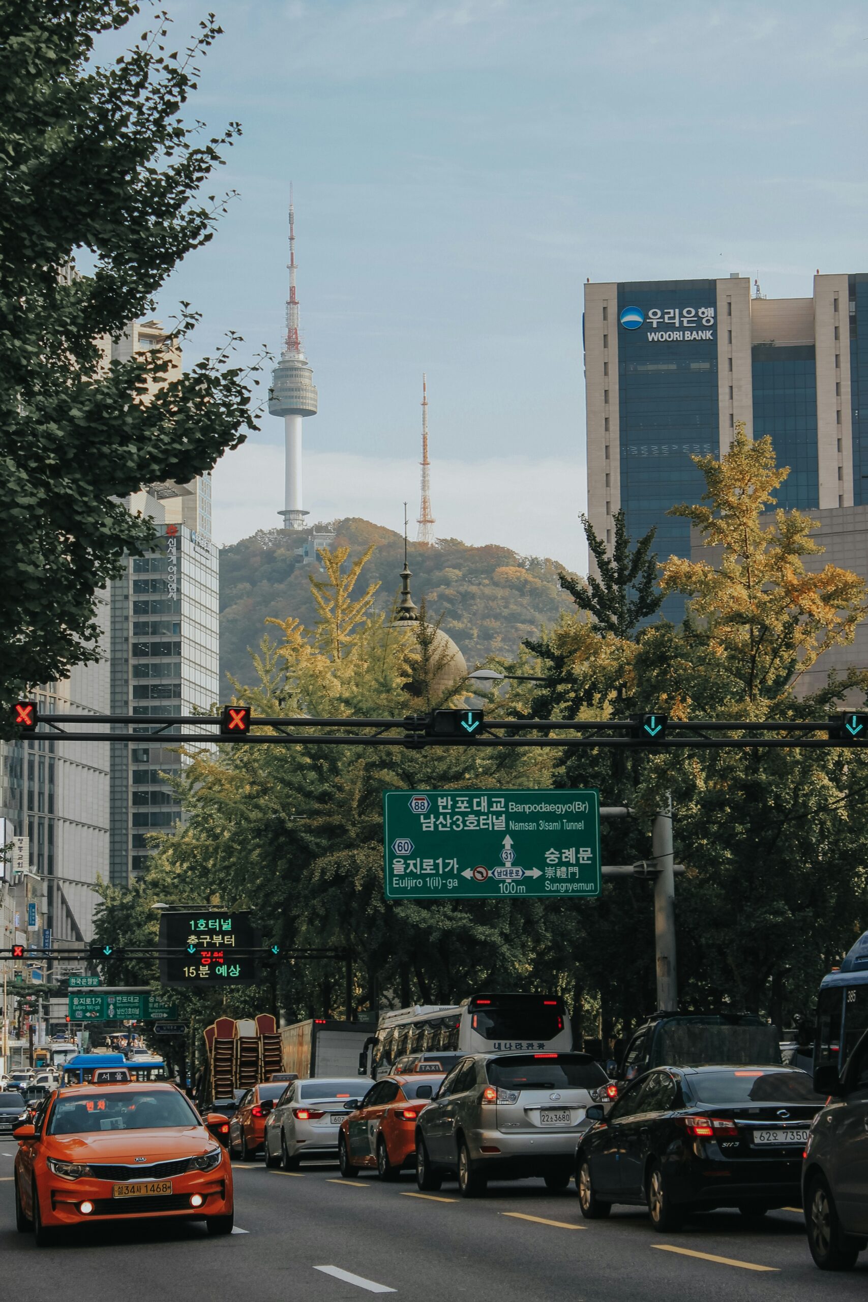 The image captures a busy street in Seoul, South Korea, with traffic, green road signs, and the iconic N Seoul Tower