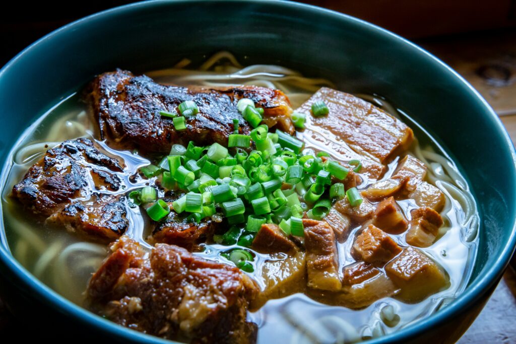 A close-up of a bowl of ramen with tender braised pork, grilled meat, and chopped green onions in a rich broth.