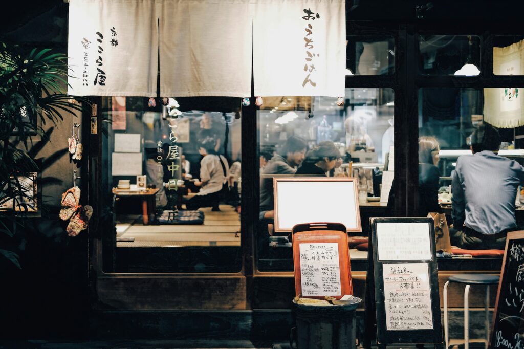 A cozy Japanese restaurant with a traditional noren curtain at the entrance, warmly lit interior, and people enjoying their meals inside